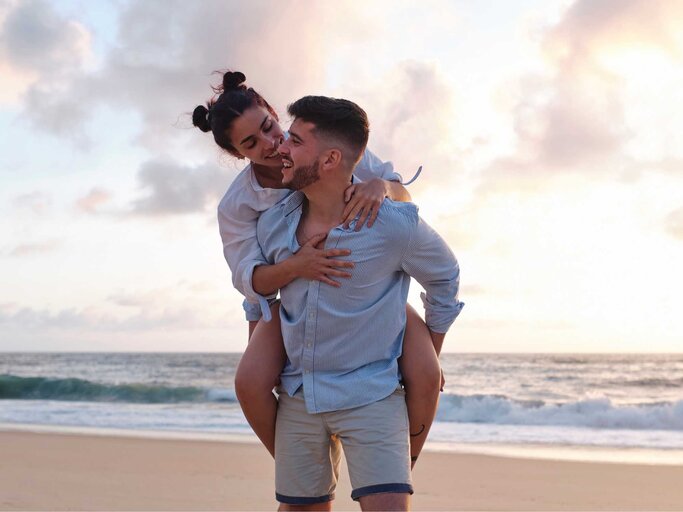 Mann trägt Partnerin Huckepack auf dem Strand | © Getty Images/Westend61