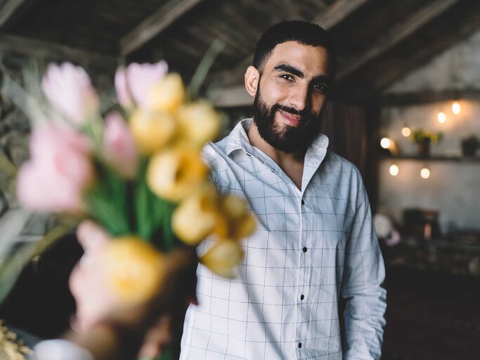 Ein Mann mit schwarzen Haaren steht im Zimmer und hält Blumen in die Kamera. | © Adobe Stock/BullRun