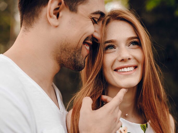 Ein Mann in weißem T-Shirt neigt sich lächelnd zu einer glücklich blickenden Frau mit langen roten Haaren und streichelt ihr Gesicht. | © Adobe Stock/Strelciuc