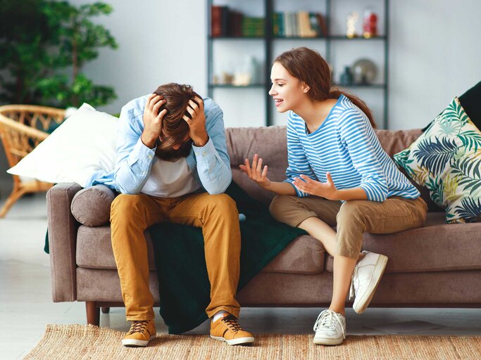 Eine junge Frau in gestreiftem Shirt gestikuliert diskutierend auf einem Sofa, während ein Mann genervt mit gesenktem Kopf seine Hände hält. | © Adobe Stock/JenkoAtaman