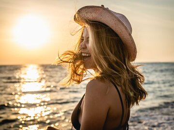 Frau lacht am Strand bei Sonnenuntergang | © Getty Images/StudioMikara