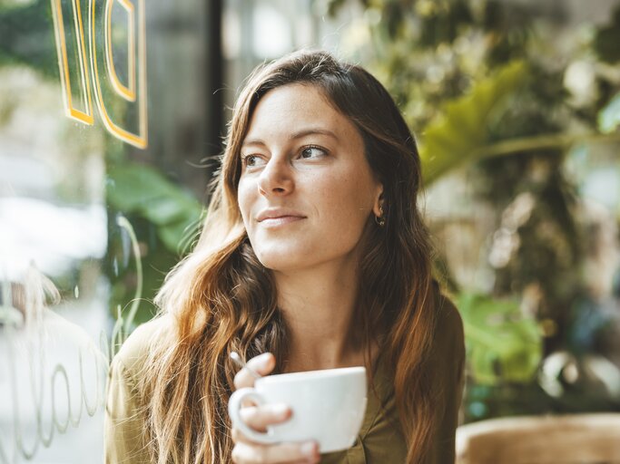 Frau mit Kaffeetasse in der Hand sitzt im Café und schaut nachdenklich aus dem Fenster | © Getty Images/Westend61