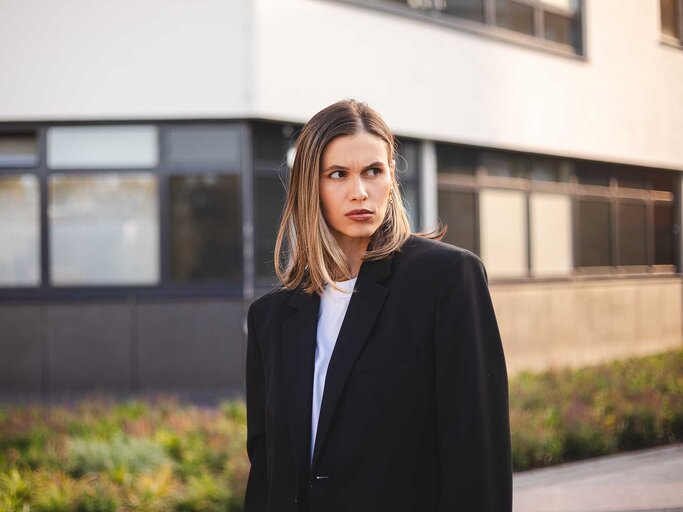 Eine Frau mit blondem Haar, schwarzem Blazer und weißem Shirt schaut skeptisch zur Seite, im Hintergrund Fenster und Grünpflanzen. | © Adobe Stock/zvkate