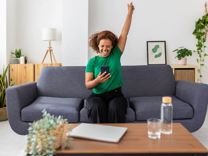 Person mit Handy in der Hand freut sich auf Sofa | © Getty Images/Xavier Lorenzo