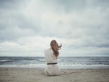 Frau sitzt im Sturm am Stand und schaut aufs Wasser. | © Adobe Stock/Aliaksei Lasevich
