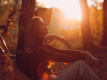 Frau sitzt im Wald an einen Baum gelehnt und die Sonne geht hinter ihr unter. Es ist Herbst. | © Adobe Stock/primipil