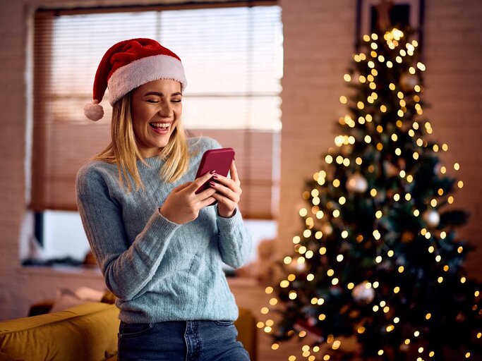 Frau trägt eine rote Nikolausmütze und steht vor einem leuchtenden Weihnachtsbaum und schaut glücklich in ihr Handy. | © Adobe Stock/Drazen