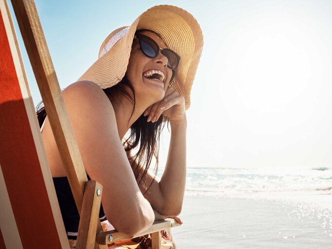 Frau sitzt im Sonnenstuhl am Strand, trägt Sonnenhut und Sonnenbrille und freut sich. | © Adobe Stock/Grady Reese/peopleimages.com