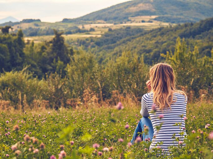 Una mujer con vaqueros y camisa a rayas está sentada sola en un prado y mira tristemente a lo lejos.  |  © Adobe Stock/AstroSystem