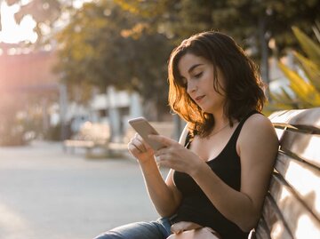 Frau sitzt auf einer Parkbank und schaut auf ihr Handy.  | © Getty Images / Cristian Blázquez Martínez