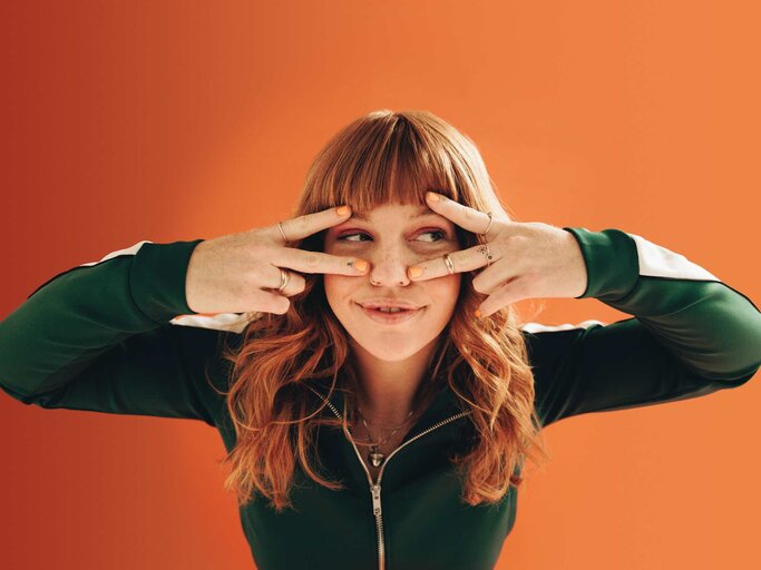 Junge Frau mit Sommersprossen und roten Haaren, die mit den Händen ein V vor ihr Gesicht hält, vor orangefarbenem Hintergrund. | © Getty Images/jacoblund