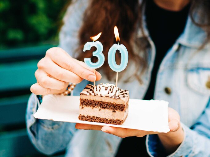 Frau hält ein Tortenstück in der Hand mit zwei Kerzen in Form der Nummer 30 | © Getty Images/Olena Malik