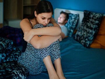 Traurige junge Frau sitzt auf ihrem Bett mit ihrem Partner im Hintergrund | © Getty Images/zoranm