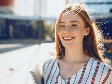 Nahaufnahme-Porträt einer jungen Frau mit roten Haaren und Sommersprossen, die in die Kamera lächelt und bei Sonnenuntergang einen Laptop hält. | © Adobe Stock/Strelciuc