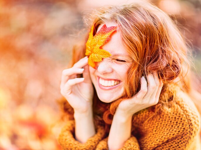 Schöne, junge Frau hält sich ein Herbstblatt vors Gesicht | © AdobeStock/drubig-photo