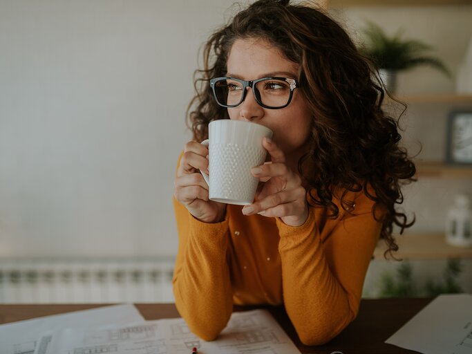 Frau mit Brille und orangem Pullover sitzt am Schreibtisch und trinkt Kaffee | © AdobeStock/Nebojsa