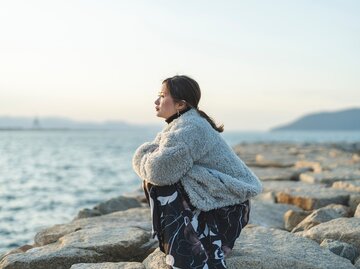 Eine Frau sitzt in der Hocke und schaut nachdenklich aufs Wasser. | © AdobeStock/peach100