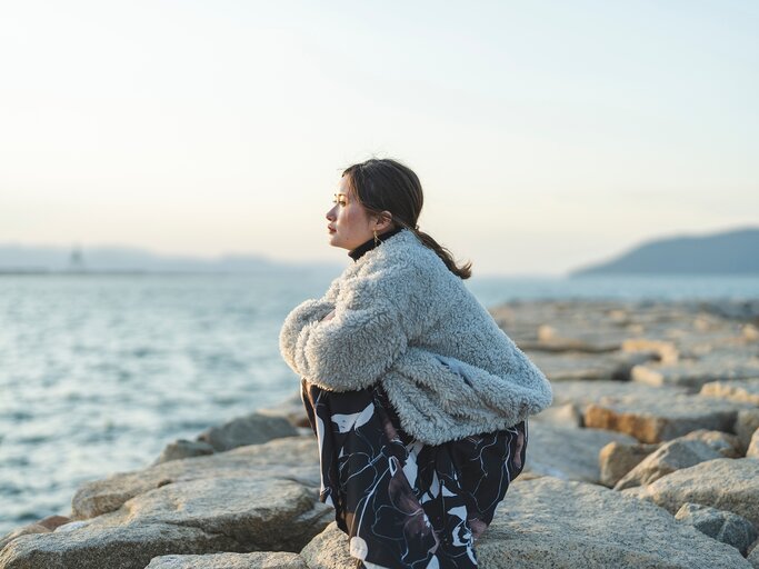 Eine Frau sitzt in der Hocke und schaut nachdenklich aufs Wasser. | © AdobeStock/peach100