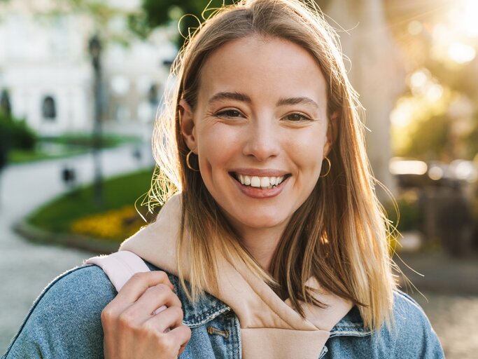 Junge Frau in Jeansjacke und Hoodie steht im Gegenlicht und lächelt in die Kamera | © AdobeStock/Drobot Dean