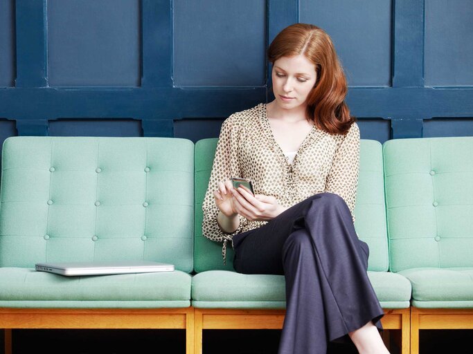 Frau sitzt auf einer Couch, hat die Beine überkreuzt und schaut in ihr Handy. | © Getty Images / Tim Robberts