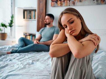 Frau sitzt unglücklich auf dem Bett, ihr Freund sitzt im Hintergrund | © Adobe Stock/HBS