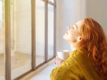 Lächelnde Frau schaut aus dem Fenster und hält ihr Gesicht in die Sonne | © Adobe Stock/contrastwerkstatt