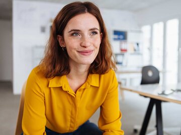 Eine junge Frau mit gelber Bluse sitzt entspannt in einem offenen Büro mit großen Fenstern und blickt glücklich zur Seite. | © Adobe Stock/contrastwerkstatt