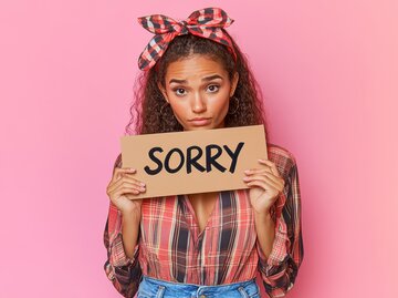 Junge Frau in kariertem Hemd hält Schild mit der Aufschrift "Sorry" in der Hand | © AdobeStock/Tuyres/Mit KI generiert