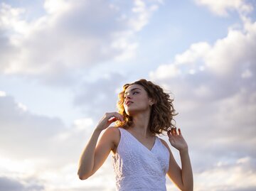 Sympathische Frau mit lockigen Haaren vor einem Wolkenhimmel | © GettyImages/Cavan Images