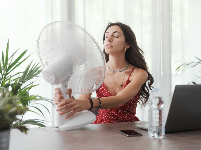 Junge Frau in rotem Kleid sitzt mit geschlossenen Augen am Tisch und richtet Ventilator auf sich, im Hintergrund Pflanzen. | © Adobe Stock/StockPhotoPro