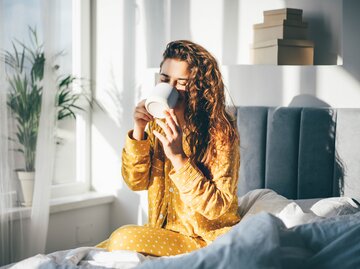 Frau sitzt im gelben Schlafanzug am Morgen im Bett und trinkt einen Kaffee | © GettyImages/Maria Korneeva