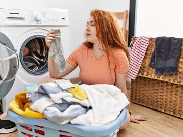 Frau sitzt vor der Waschmaschine und hält eine Socke in die Luft | © Adobe Stock/Krakenimages.com