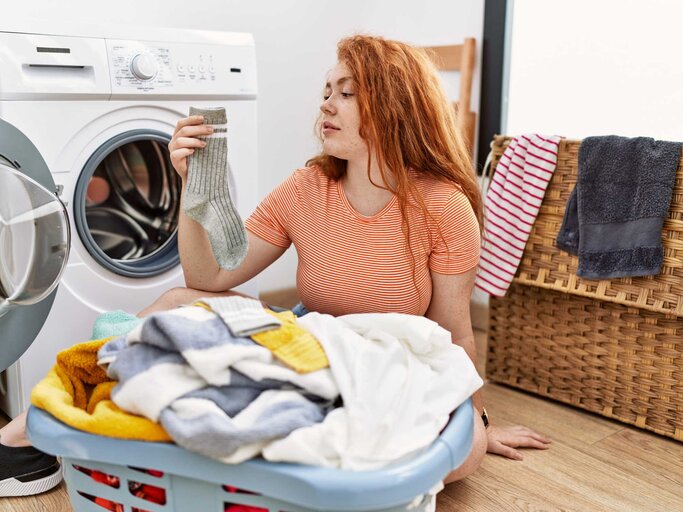 Frau sitzt vor der Waschmaschine und hält eine Socke in die Luft | © Adobe Stock/Krakenimages.com