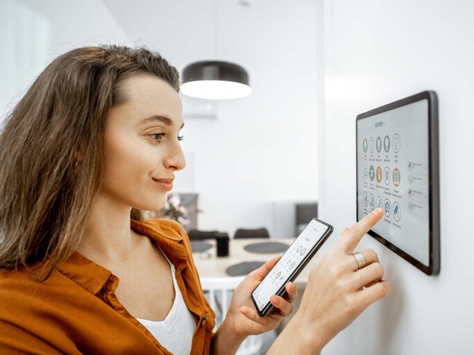 Junge Frau, die ihr Haus mit einem digitalen Touchscreen-Panel an der Wand im Wohnzimmer steuert.  | © Adobe Stock/rh2010