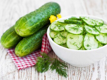 Eine Schüssel mit Gurkensalat steht auf einem Tisch mit rot-weiß karierter Tischdecke. Daneben liegen ein Bund Bill und Gurken. | © Getty Images / Dorin Vladu / 500px