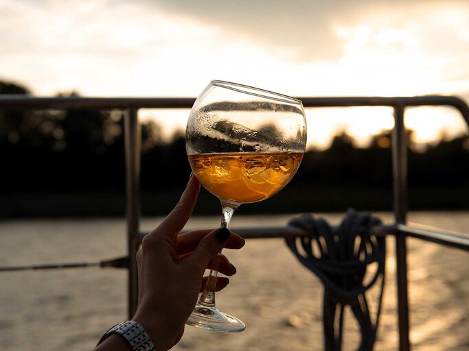 Hand einer Frau hält ein Glas mit Eiswürfeln und einem Aperol Getränk in den Sonnenuntergang. | © Adobe Stock/Fostic