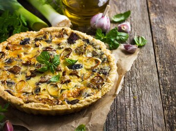 Eine Quiche auf einem Holzbrett umgeben von Gemüse | © Adobe Stock/anna_shepulova