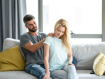 Mann massiert seiner Frau im Wohnzimmer die Schultern. | © Getty Images/DjelicS