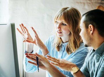 Frau und Mann sitzen vor einem Computerbildschirm und sprechen über ihre Arbeit. | © Getty Images / Hinterhaus Productions