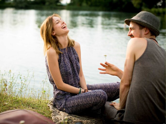 Frau und Mann sitzen am See und lachen zusammen | © Getty Images/Oana Szekely