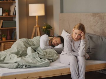 Person sitzt besorgt auf Bett neben schlafendem Partner | © Getty Images/stockphotodirectors