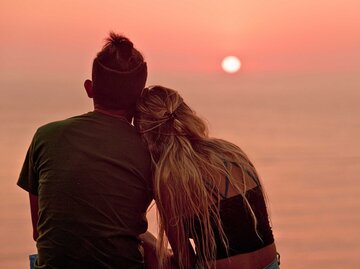 Silhouette eines Paares, bestehend aus einer blonden Frau mit Pferdeschwanz, die auf der Schulter eines Mannes mit Pferdeschwanz ruht, der den Sonnenuntergang in Barranco, Lima, Peru, beobachtet. | © Adobe Stock/Franz