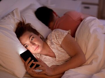 Frau liegt mit ihrem Partner im Bett und schaut auf ihr Handy | © Adobe Stock/Syda Productions