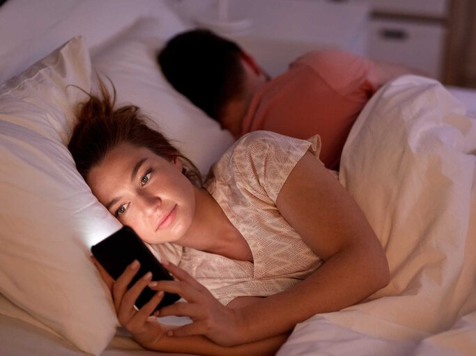 Frau liegt mit ihrem Partner im Bett und schaut auf ihr Handy | © Adobe Stock/Syda Productions