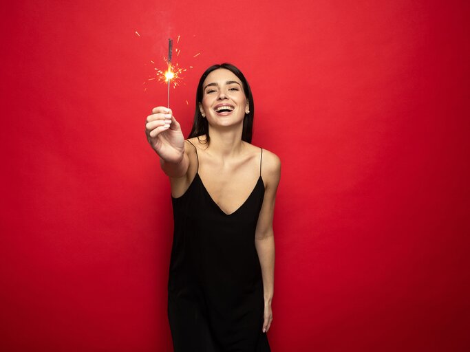 Junge Frau in schwarzem Slipdress steht vor rotem Hintergrund und hält Wunderkerze in Hand | © AdobeStock/makedonski2015