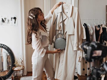 Frau steht in einem hellen Zimmer und einen einen Kleiderbügel, an dem ein beiger Trenchcoat hängt in die Luft und betrachtet diesen. | © Adobe Stock/Look!