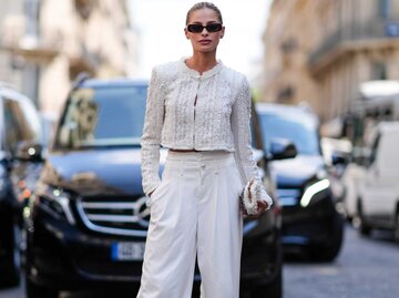 Eine Frau trägt eine schwarze Sonnenbrille, weiße gemusterte kurze Jacke, weiße weite Hosen, weiße Paillettentasche und hellbraune Lederschuhe. | © Getty Images/Edward Berthelot 