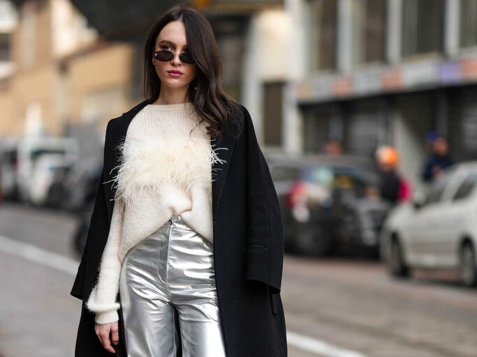 Frau trägt schwarze Sonnenbrille, weißen Wollpullover, schwarzen Mantel und weite silberne Hosen  | © Getty Images/Edward Berthelot 