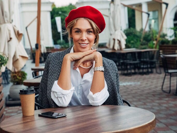 Frau sitzt an Café-Tisch mit Baskenmütze | © Getty Images/CoffeeAndMilk