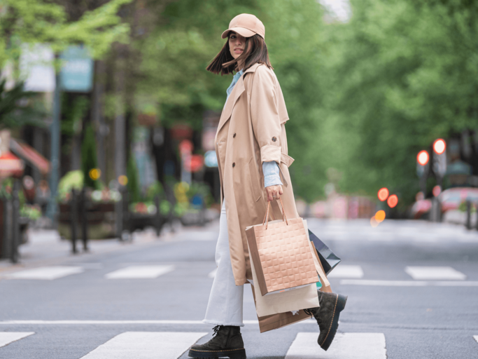 Frau mit Tüten in Trenchcoat überquert Straße | © Getty Images/Westend61